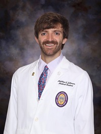 Professional medical school portrait of a person wearing a white medical coat, white dress shirt, and patterned tie against a dark blue-gray studio background.