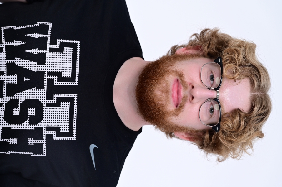 Head-and-shoulders portrait of a person with curly hair, glasses, and a beard wearing a black graphic T-shirt against a plain light background.