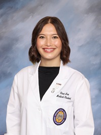 Professional portrait of a person wearing a white medical coat over a black top against a soft blue studio background.