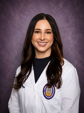 Professional medical school portrait of a person wearing a white medical coat over a dark top against a dark purple studio background.
