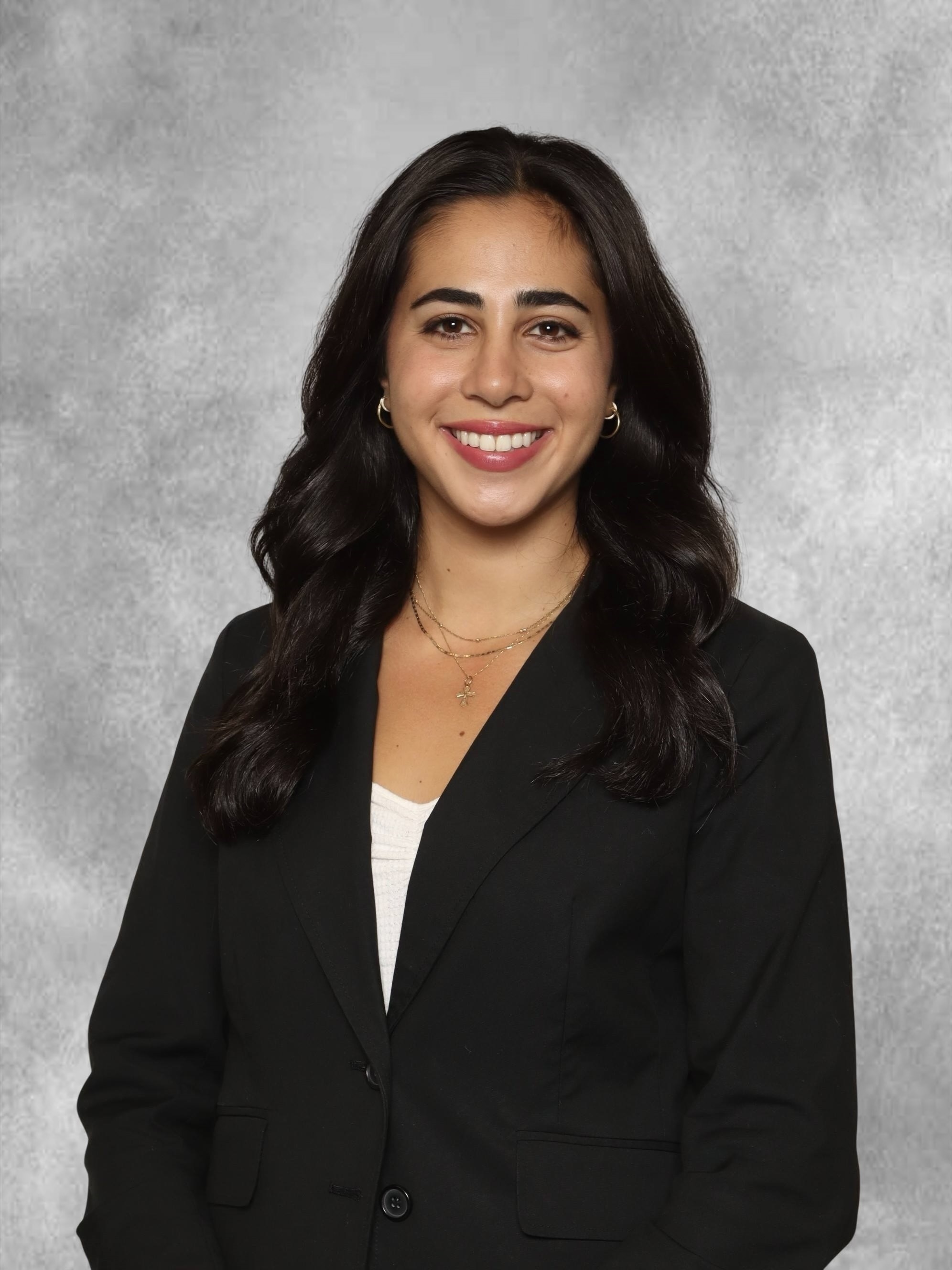 Professional portrait of a person wearing a black blazer over a light-colored top against a gray studio background.