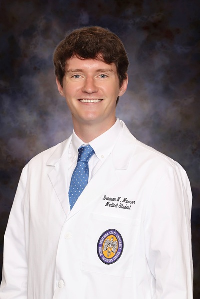 Professional medical school portrait of a person wearing a white medical coat, white dress shirt, and light blue tie against a dark studio background.