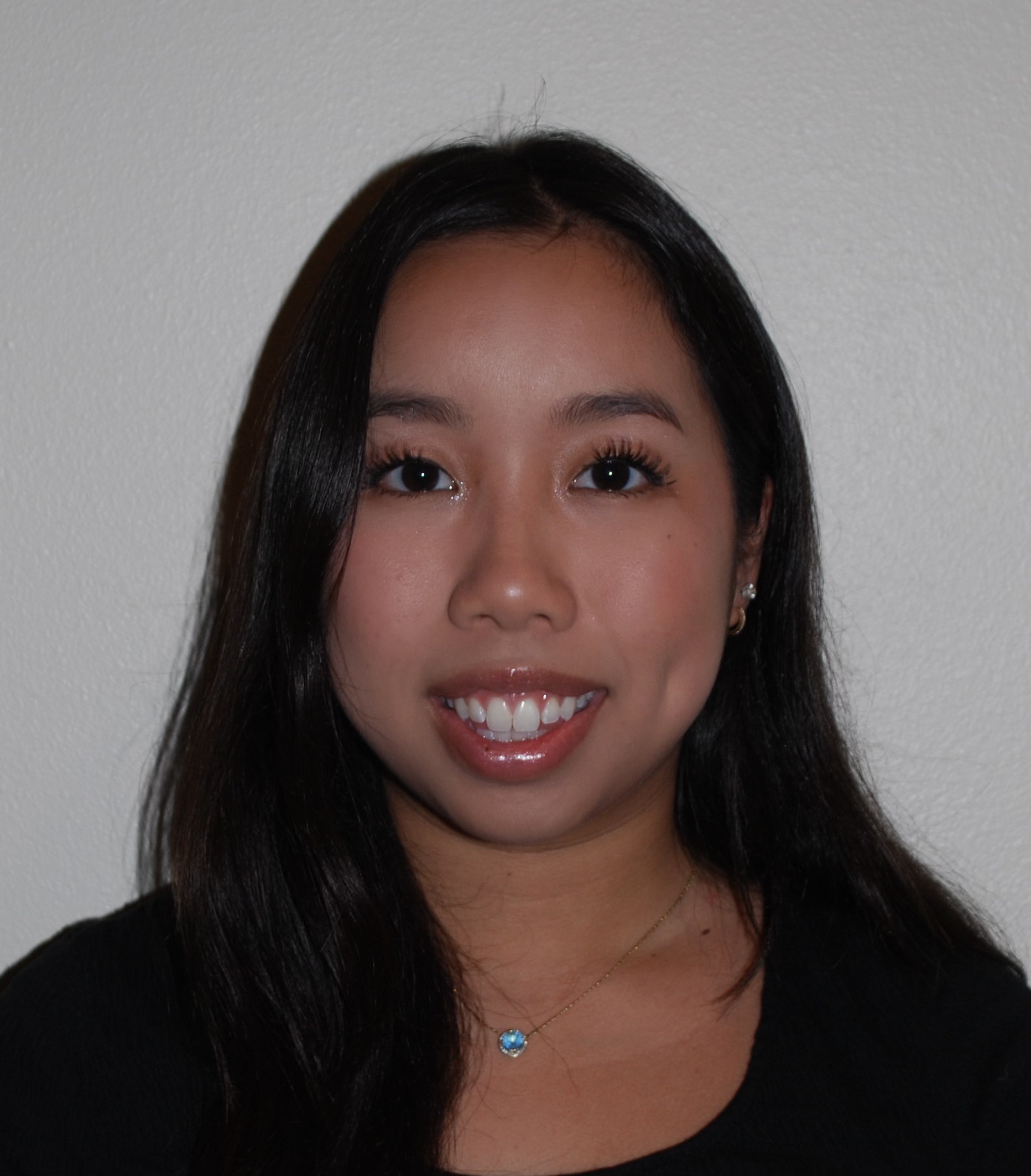 Professional headshot of a person with long dark hair wearing a black top and a small pendant necklace against a plain light-colored background.
