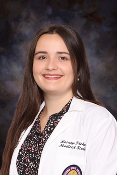 Professional medical school portrait of a person wearing a white medical coat over a patterned dark blouse against a dark blue-gray studio background.