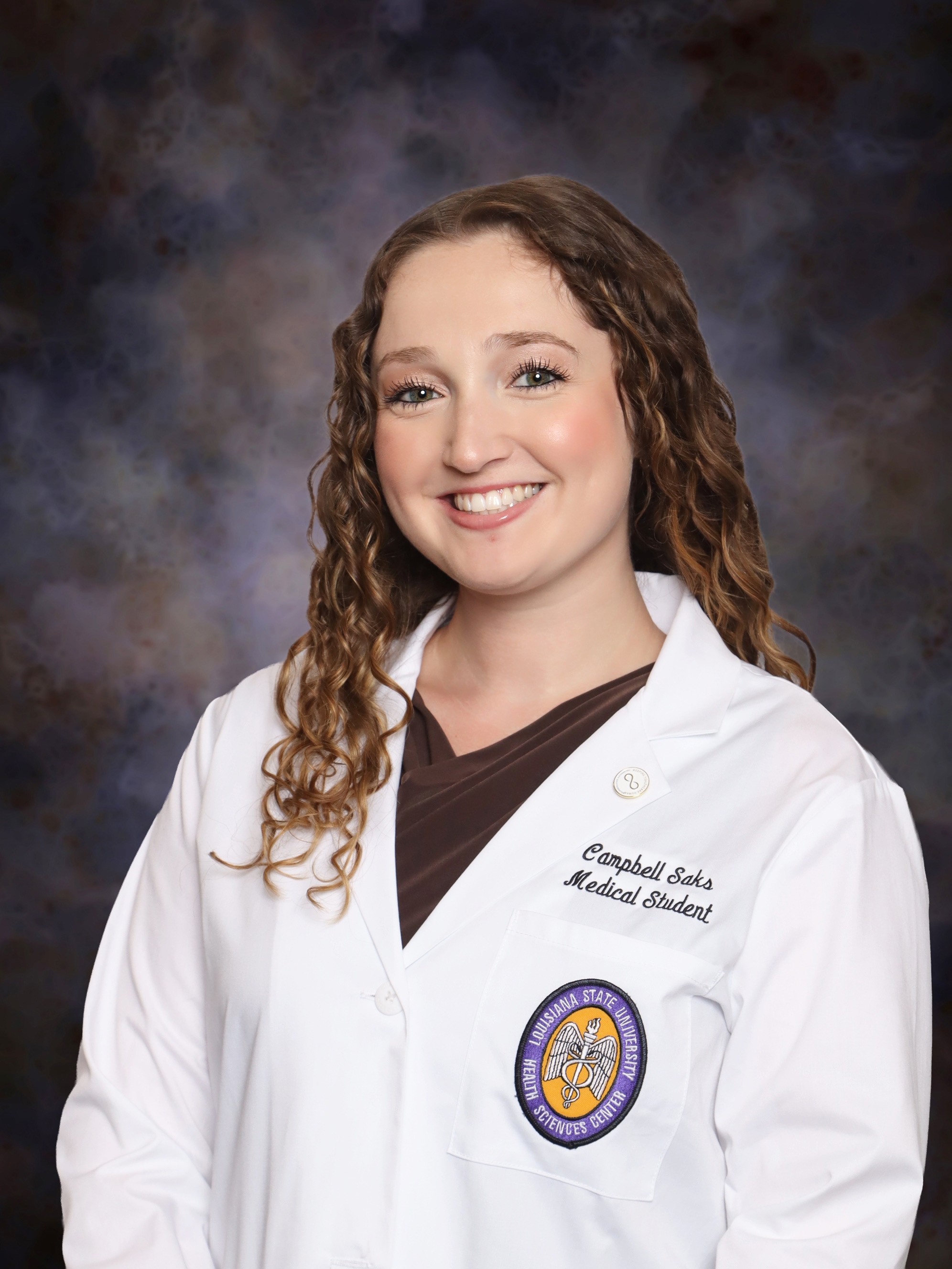 Professional medical school portrait of a person wearing a white medical coat over a dark brown top against a dark blue-gray studio background.