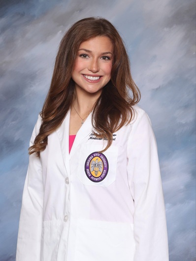 Professional portrait of a person wearing a white medical coat over a bright pink top against a light blue studio background.