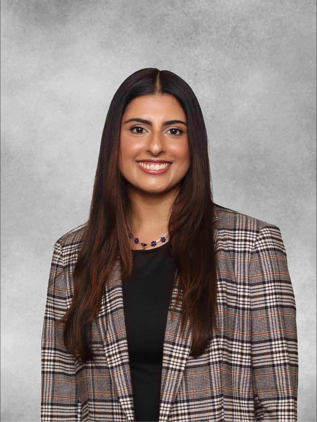 Professional headshot showing a person wearing a plaid blazer over a dark top and a beaded necklace. The subject has long hair and is facing the camera against a light gray studio background. The image is evenly lit and presented in a formal portrait style.
