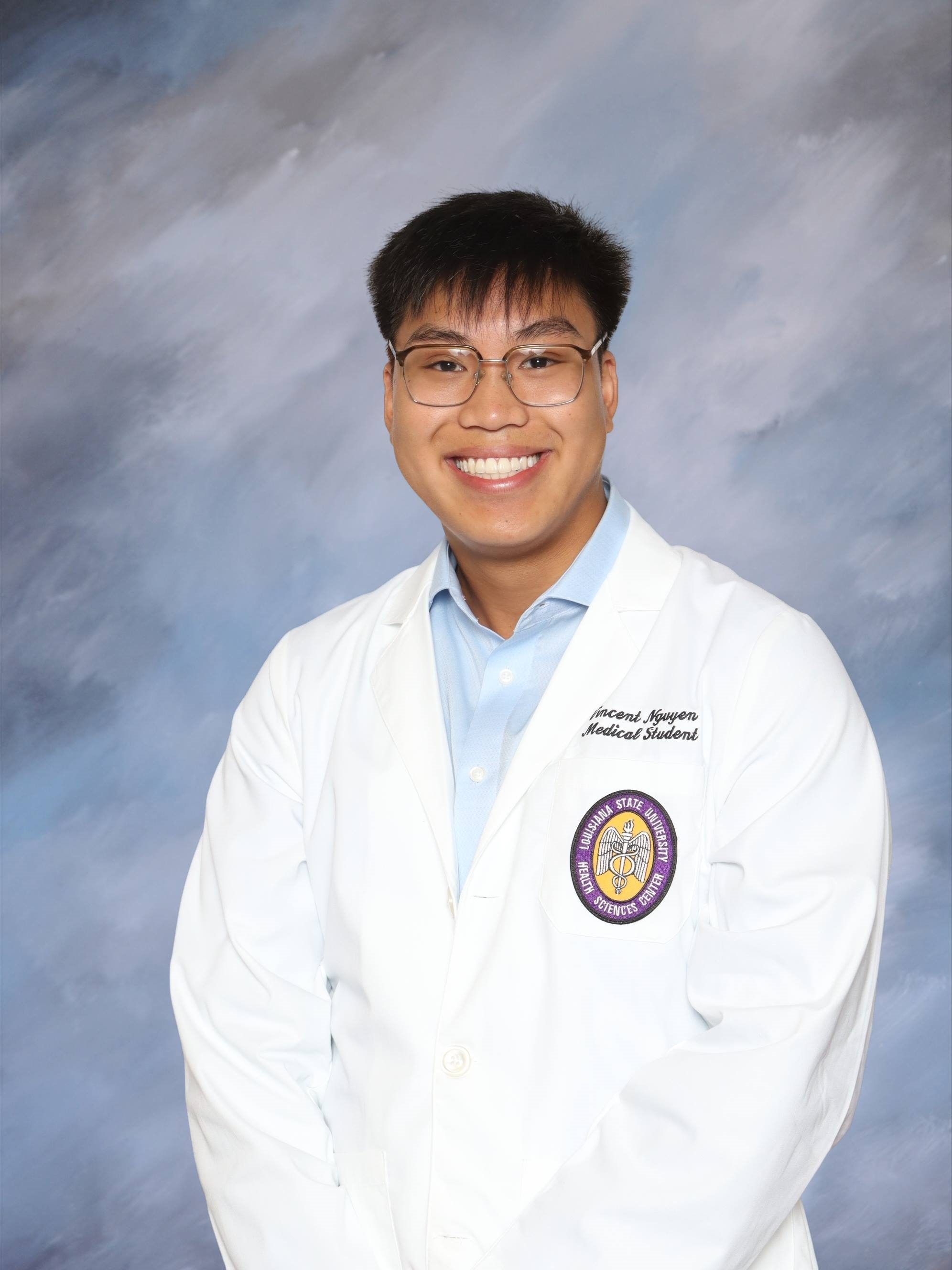 Professional medical school portrait showing a person wearing a white medical coat over a light blue collared shirt. Visible embroidered text on the coat includes the title “Medical Student,” and a Louisiana State University Health Sciences Center emblem is displayed on the chest pocket. The subject is wearing rectangular eyeglasses and is facing the camera against a softly textured light blue and gray studio background. The image is evenly lit and presented in a formal portrait style.