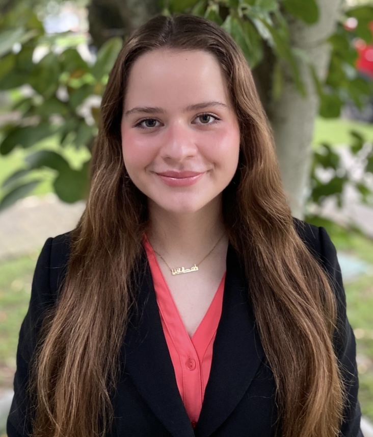 Professional outdoor headshot showing a person wearing a black blazer over a coral-colored blouse. The subject has long hair and is wearing a necklace. The portrait is taken outdoors in natural daylight, with a tree trunk and green foliage visible in the background. The image is evenly focused on the subject with a softly blurred background.