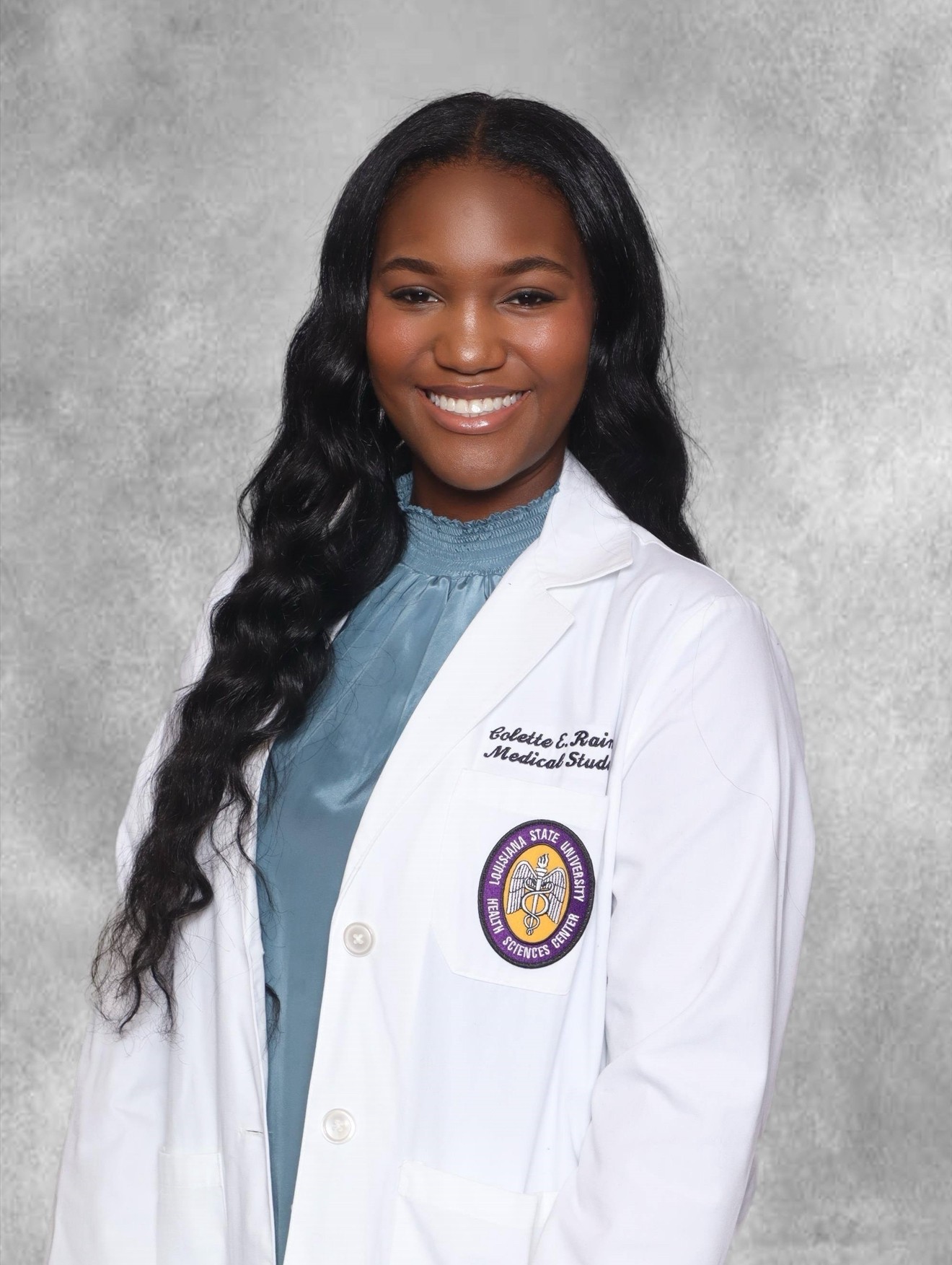 Professional headshot of a person standing against a light gray textured studio background. The person has long dark wavy hair and is wearing a white medical student coat over a light blue high-neck blouse. An embroidered patch for Louisiana State University Health Sciences Center is visible on the left chest pocket of the coat. The image is framed from the upper thighs up, with the person facing the camera in a formal portrait pose.