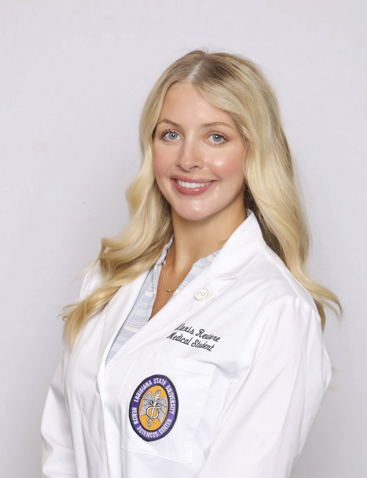 Professional medical school headshot showing a person wearing a white medical coat with embroidered text identifying the wearer as a medical student and a Louisiana State University Health Sciences Center emblem on the chest pocket. The subject is wearing a light-colored striped top and a small necklace beneath the coat. A lapel pin is attached to the coat. The portrait is set against a plain light gray background with even, professional lighting.