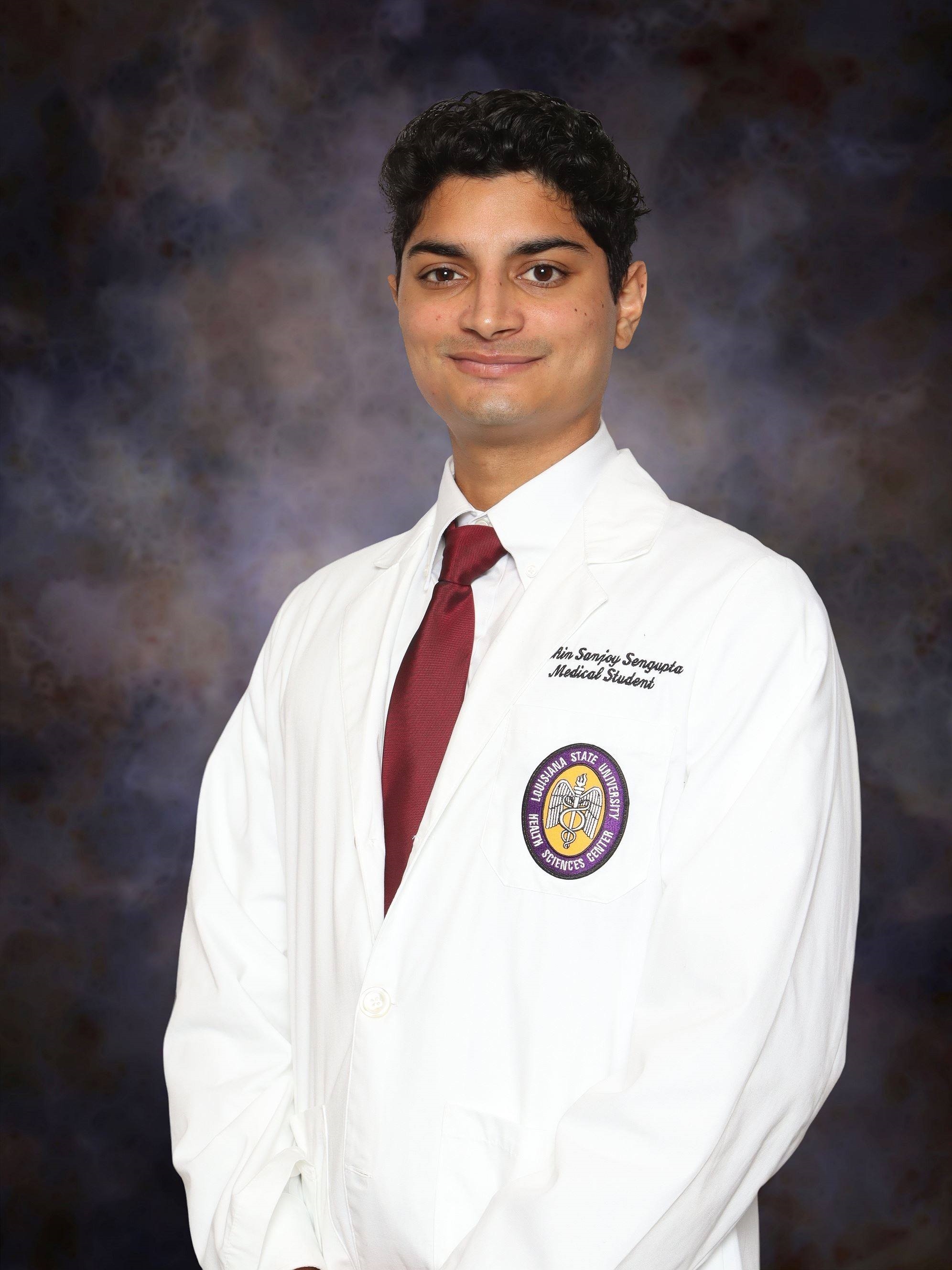 Professional studio headshot of a person standing against a dark, mottled blue and gray backdrop. The person is wearing a white medical student coat over a white dress shirt with a burgundy tie. An embroidered Louisiana State University Health Sciences Center patch is visible on the left chest pocket of the coat. The portrait is framed from about the waist up, with the person facing slightly to one side while looking toward the camera. Visible embroidered text on the coat identifies the wearer as a medical student.