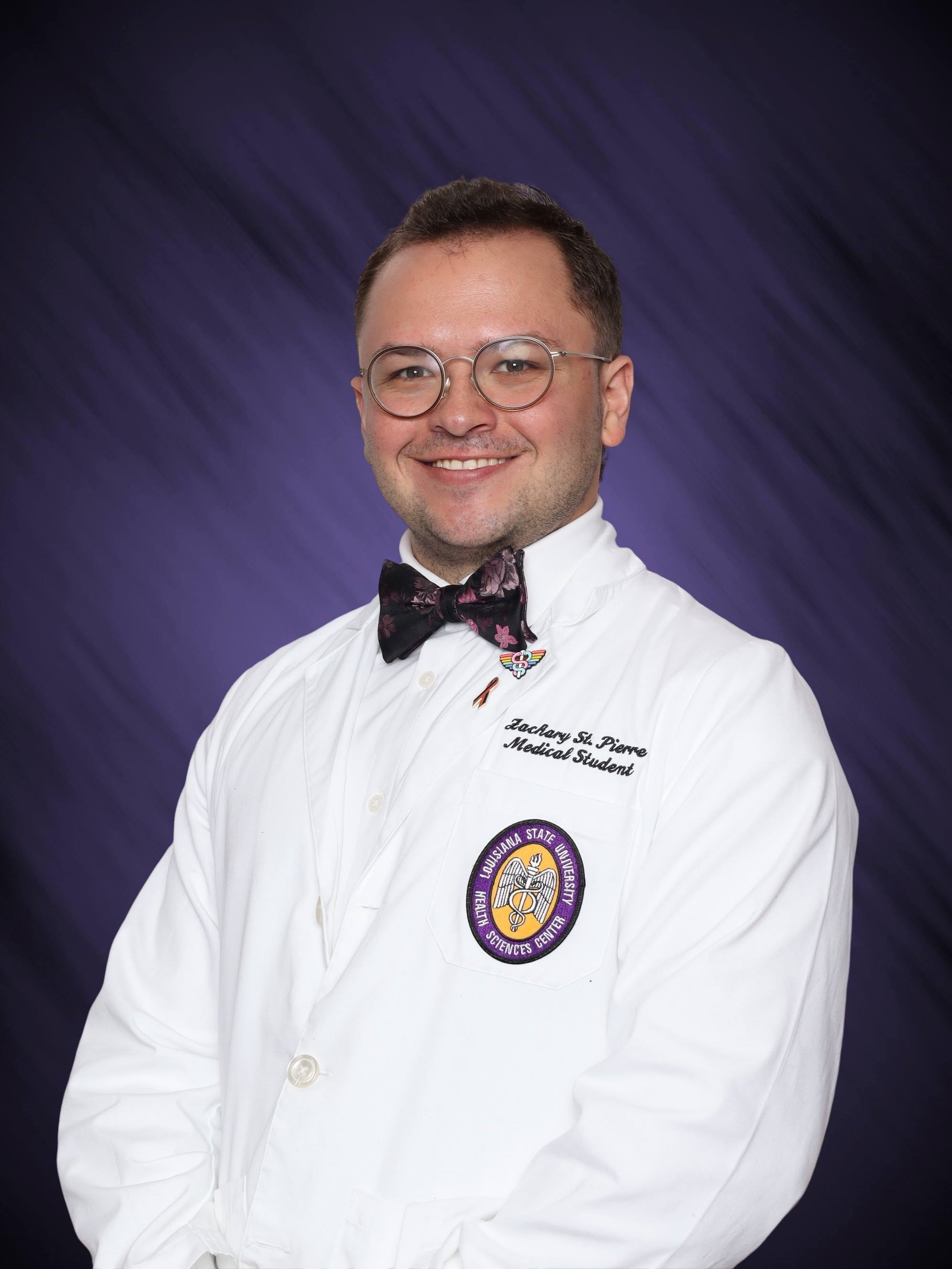 Professional medical school headshot showing a person wearing a white medical coat with embroidered text identifying the wearer as a medical student and a Louisiana State University Health Sciences Center emblem on the chest pocket. The subject is wearing round eyeglasses, a patterned bow tie, and multiple lapel pins on the coat. The portrait is set against a dark purple studio background with subtle diagonal texture and even, professional lighting.
