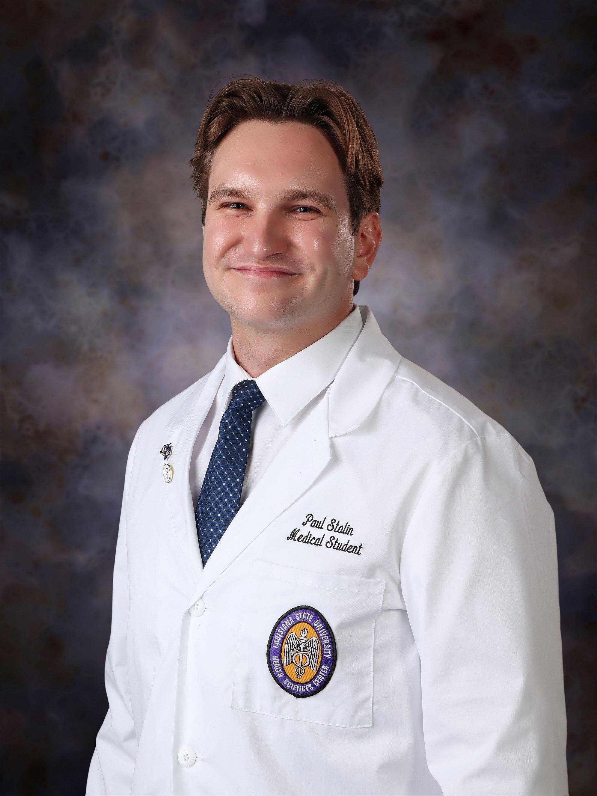 Professional medical school headshot showing a person wearing a white medical coat over a white dress shirt and blue patterned tie. Visible embroidered text on the coat includes the title “Medical Student,” and a Louisiana State University Health Sciences Center emblem is displayed on the chest pocket. Two lapel pins are attached to the coat. The subject is facing the camera against a dark blue and gray mottled studio background with even, professional lighting.