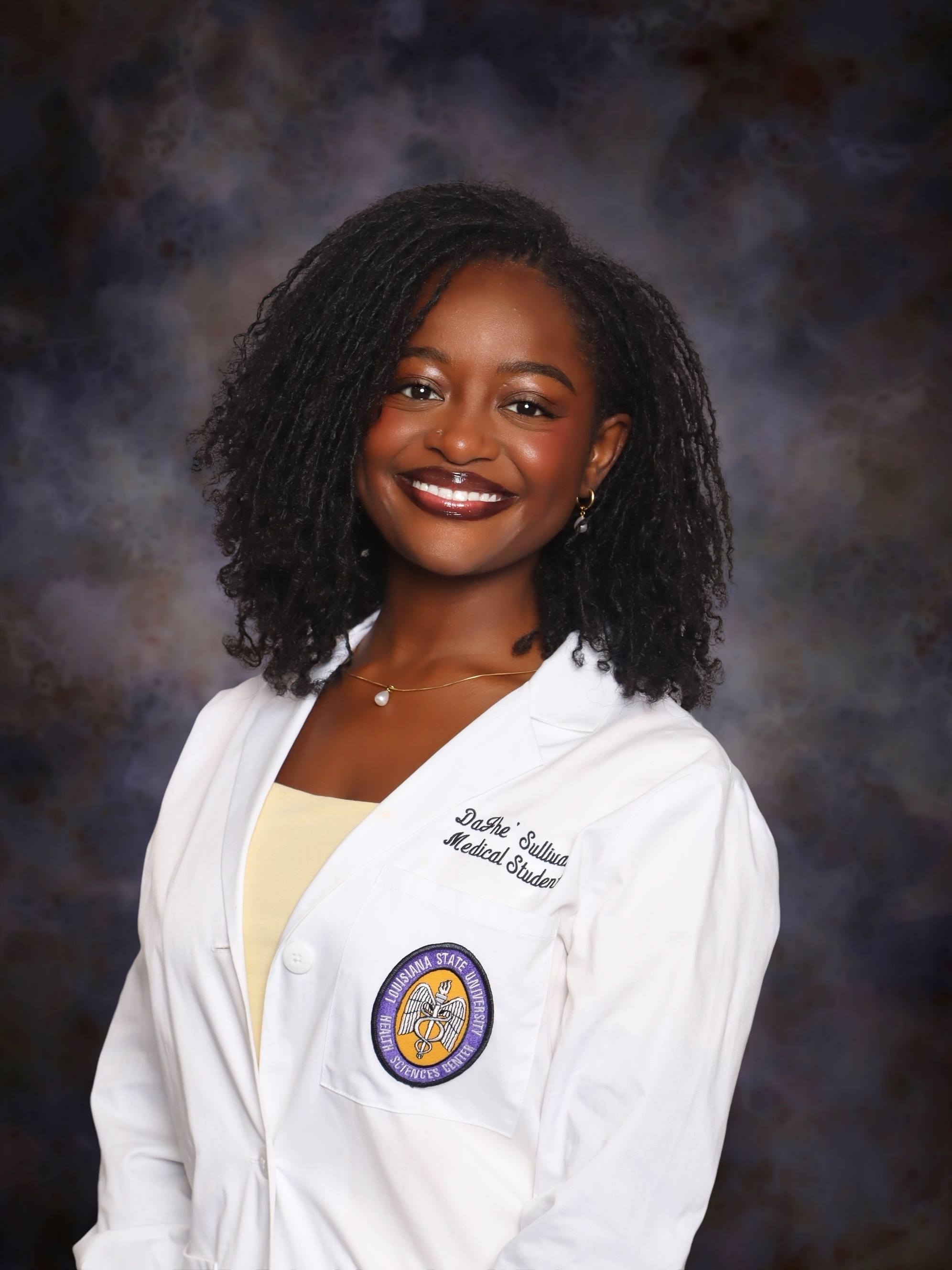 Professional medical school portrait showing a person wearing a white medical coat over a light-colored top. Visible embroidered text on the coat includes the title “Medical Student,” and a Louisiana State University Health Sciences Center emblem is displayed on the chest pocket. The subject is wearing a delicate necklace and earrings. The portrait is set against a dark blue and gray mottled studio background with even, professional lighting.