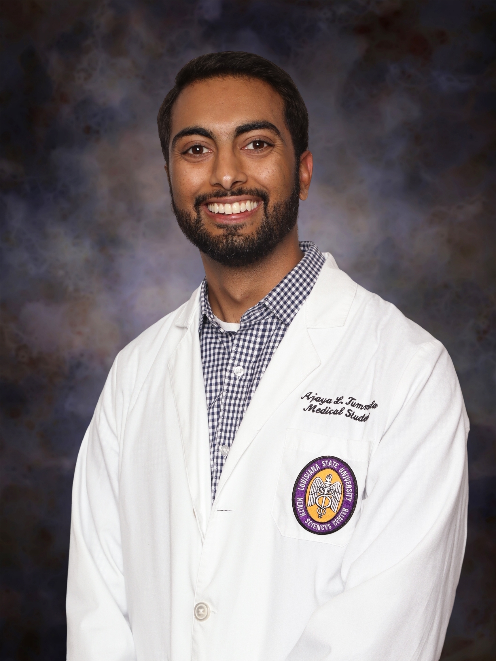 Professional studio headshot of a person standing against a dark blue and gray mottled backdrop. The person is wearing a white medical student coat over a blue-and-white checkered button-down shirt. An embroidered Louisiana State University Health Sciences Center patch is visible on the left chest pocket of the coat. The portrait is framed from about the waist up, with the person facing slightly to one side while looking toward the camera. Embroidered text on the coat identifies the wearer as a medical student.
