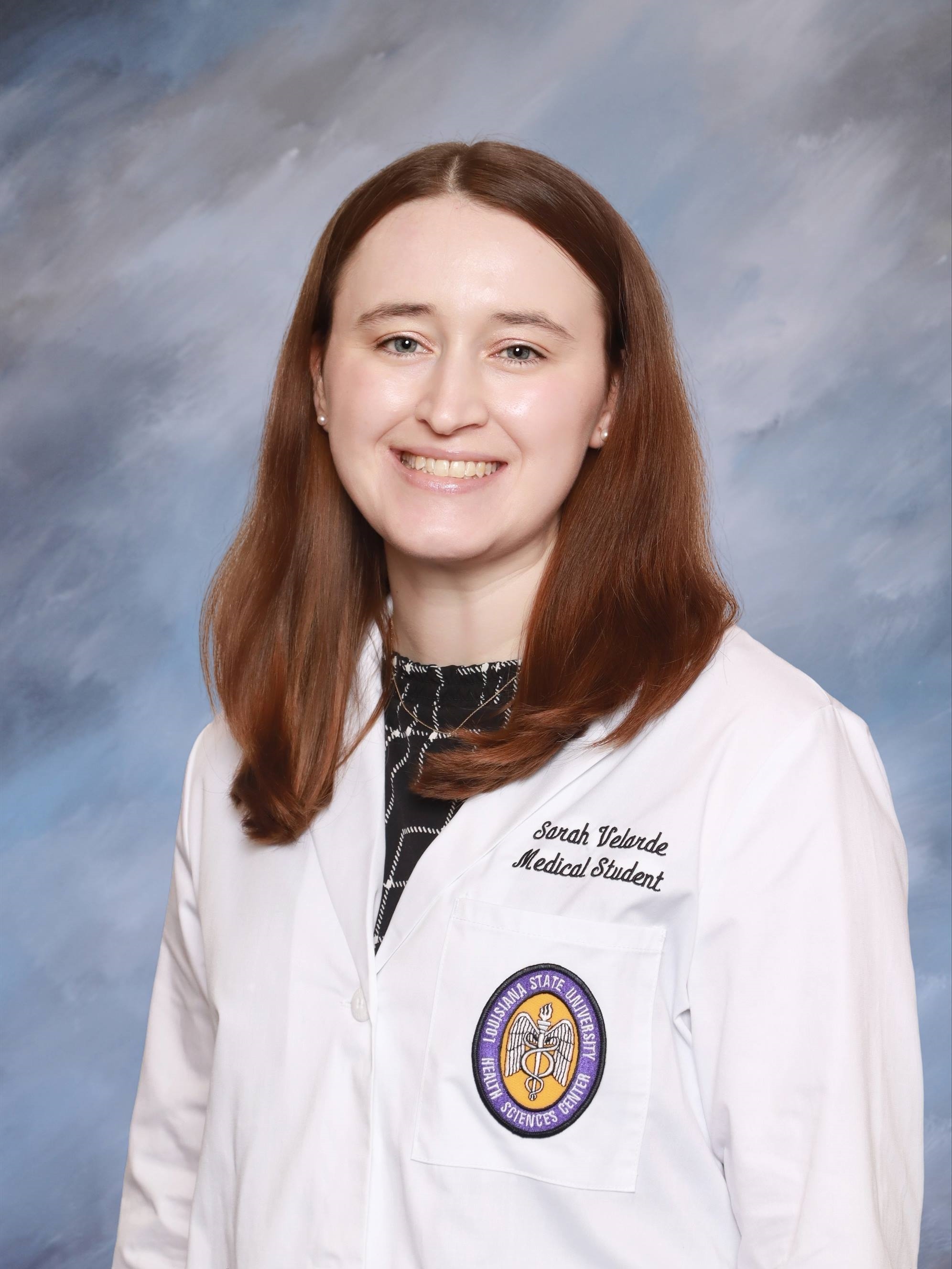 Professional medical school headshot showing a person wearing a white medical coat with embroidered text identifying the wearer as a medical student and a Louisiana State University Health Sciences Center emblem on the chest pocket. The subject is wearing a patterned dark top beneath the coat and small stud earrings. The portrait is set against a softly textured light blue studio background with even, professional lighting.