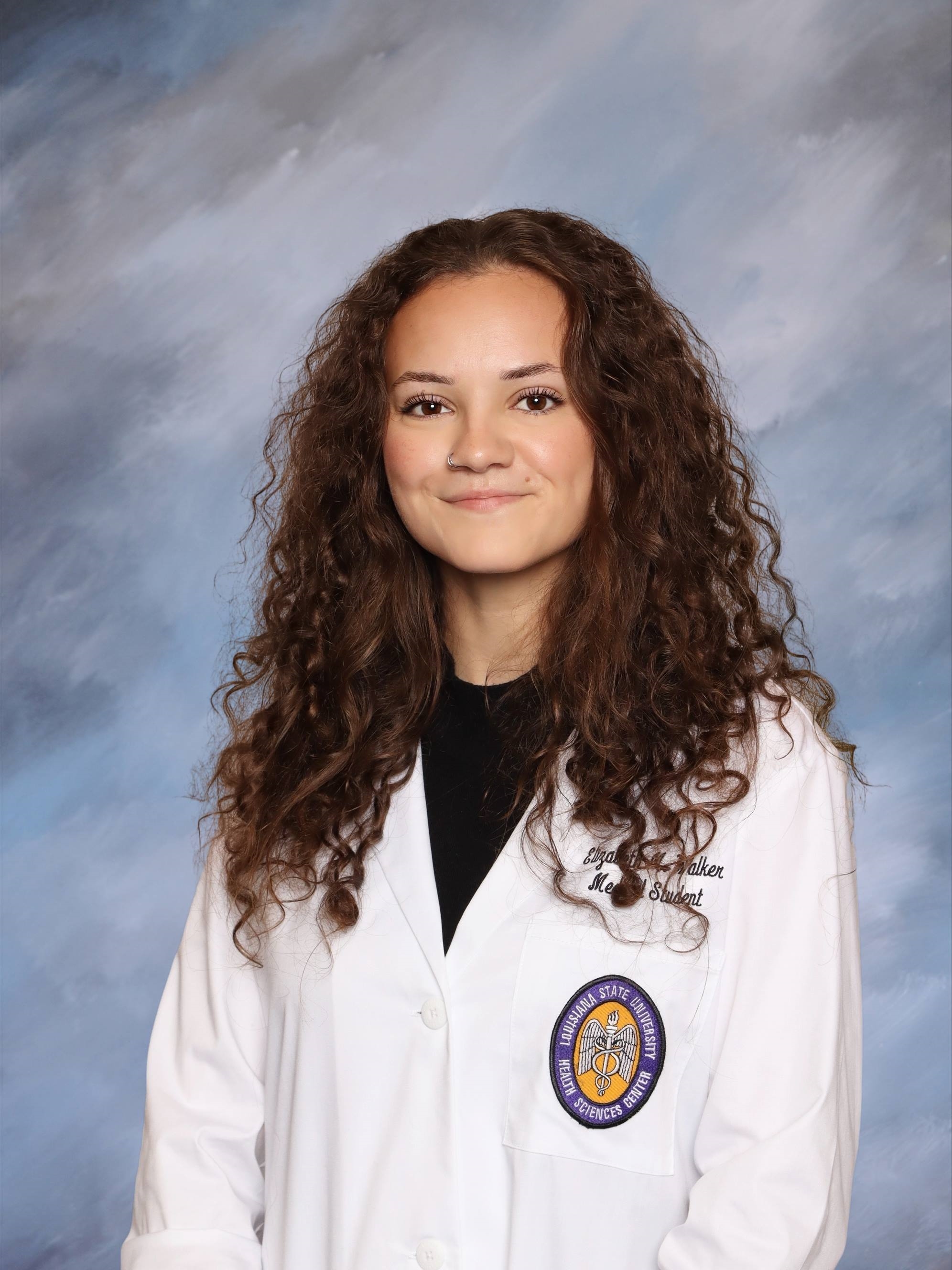 Professional medical school portrait showing a person wearing a white medical coat over a black top. Visible embroidered text on the coat includes the title “Medical Student,” and a Louisiana State University Health Sciences Center emblem is displayed on the chest pocket. The subject has long curly hair and a nose ring and is facing the camera against a softly textured light blue and gray studio background. The image is evenly lit and presented in a formal portrait style.