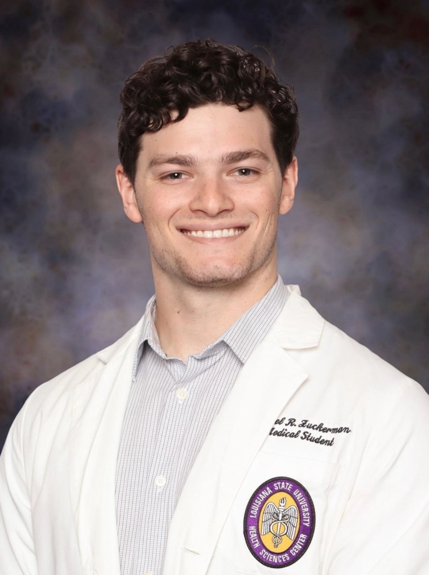Professional studio headshot of a person standing against a dark blue and gray mottled backdrop. The person is wearing a white medical student coat over a light gray checkered button-down shirt. An embroidered Louisiana State University Health Sciences Center patch is visible on the left chest pocket of the coat. The portrait is framed from the chest up, with the person angled slightly to one side while looking toward the camera. Embroidered text on the coat identifies the wearer as a medical student.