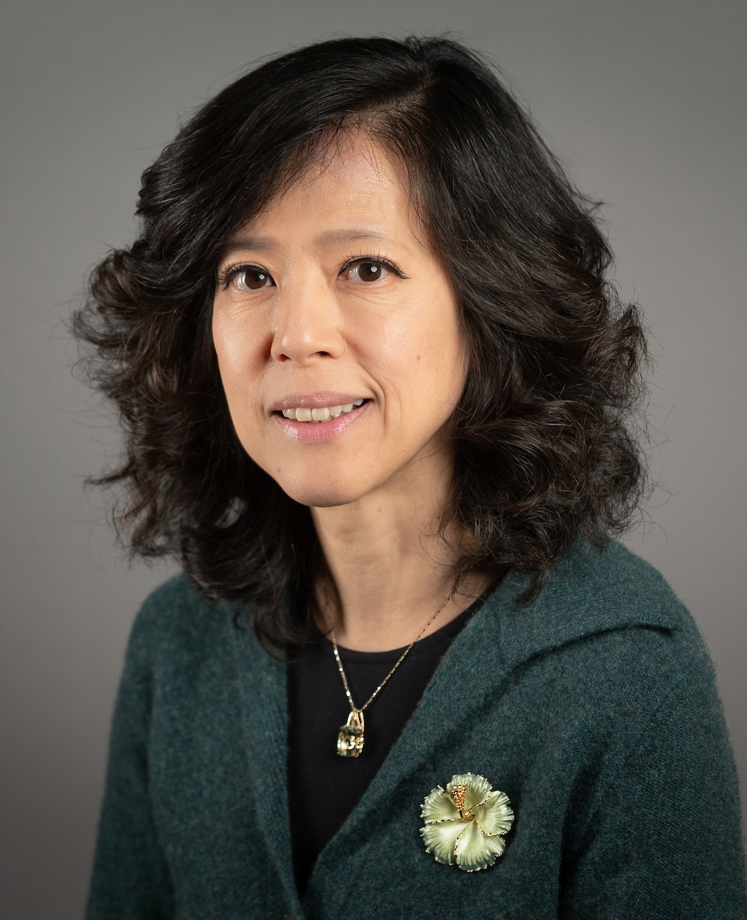 Professional headshot of a person with shoulder-length dark wavy hair, wearing a dark green cardigan over a black top. A pendant necklace and a flower-shaped brooch are visible on the cardigan. The person is facing the camera in a three-quarter pose against a neutral gray studio background. The image is evenly lit and cropped from the chest up, creating a formal portrait suitable for a professional profile.