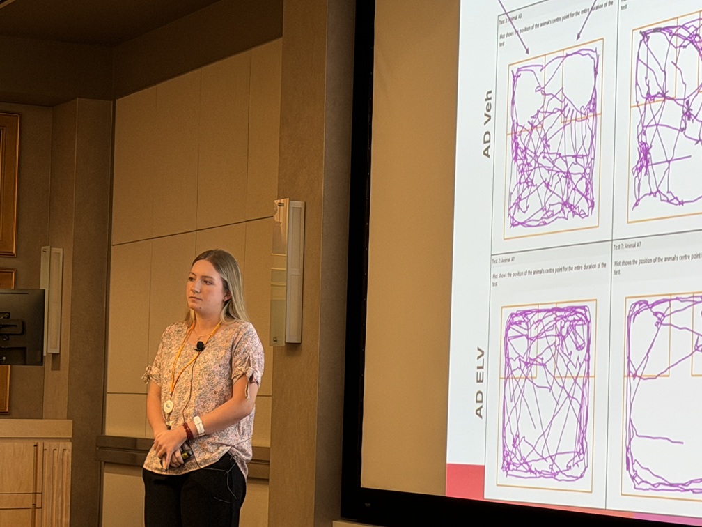 A presenter stands beside a large projection screen in a conference room, delivering a research presentation. The slide displays multiple panels of purple line-tracing plots labeled “AD Yeh” and “AD ELV,” likely illustrating visual or eye-tracking data comparisons. The presenter wears a microphone and stands near the front of the room, while the screen dominates the right side of the image. Light-colored wall panels, a lectern, and framed wall portraits are visible in the background, creating a formal academic setting.