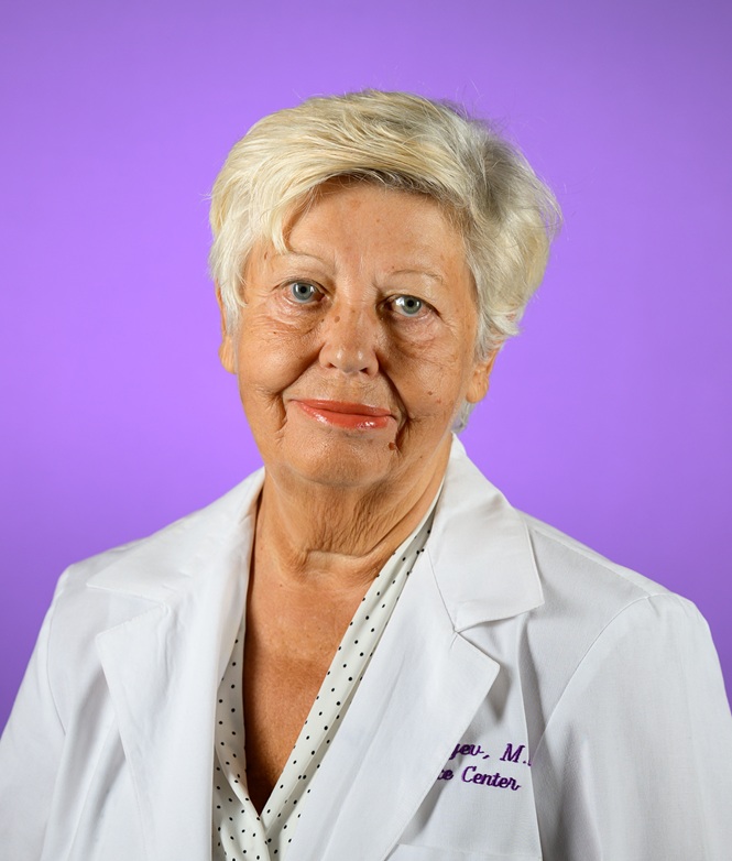 Head-and-shoulders studio portrait of a person with short, light-colored hair wearing a white lab coat and a light blouse with a subtle dotted pattern. The subject faces the camera against a smooth purple gradient background. Partial embroidered text is visible on the lab coat, though it is not fully legible. The image is evenly lit and professionally photographed.