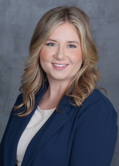 Professional headshot of a person with shoulder-length wavy hair, photographed against a gray studio backdrop. The person is wearing a navy-blue blazer over a light-colored blouse and is posed at a slight angle toward the camera. The portrait is cropped from the chest up and uses soft, even lighting typical of a business or organizational profile photo.