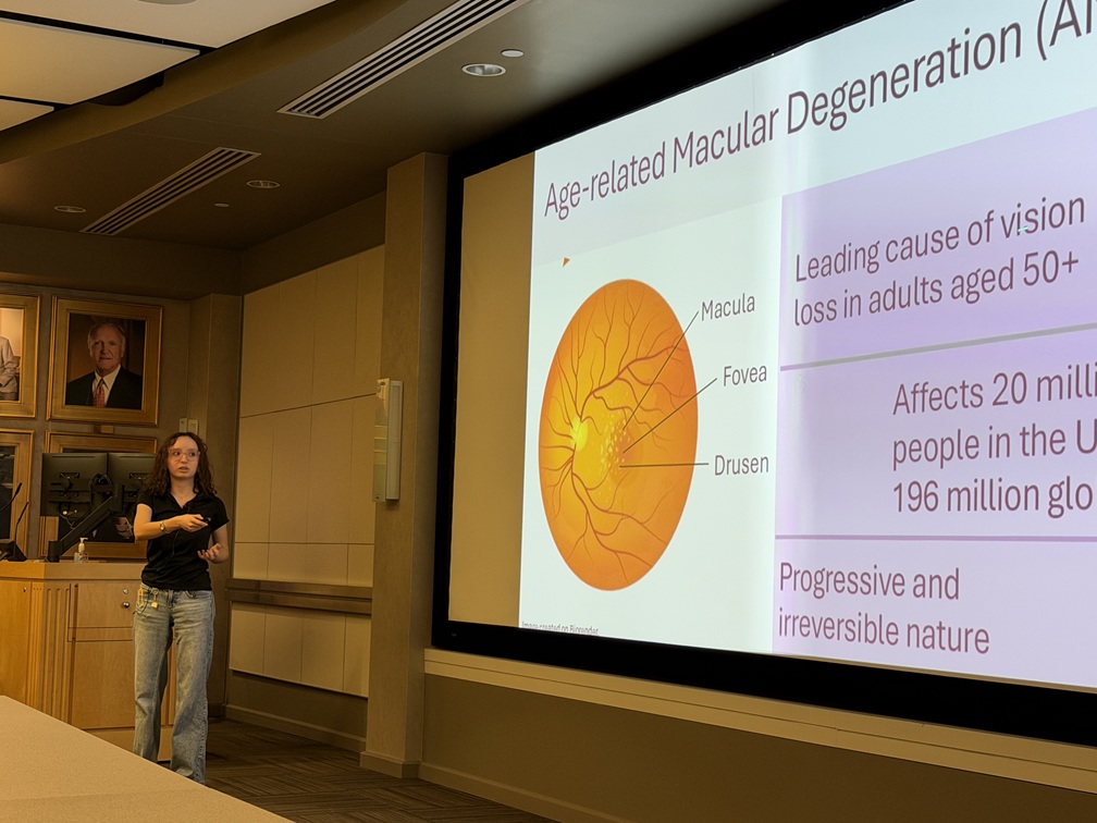 A presenter stands at the front of a conference room beside a large projection screen displaying a slide about age-related macular degeneration (AMD). The slide includes a labeled diagram of the retina identifying the macula, fovea, and drusen, along with key facts about vision loss in adults over age 50 and the prevalence of AMD. Framed portraits hang on the wall near a lectern in the background, while tables and seating are visible in the foreground. The photo captures an academic or scientific presentation in progress.