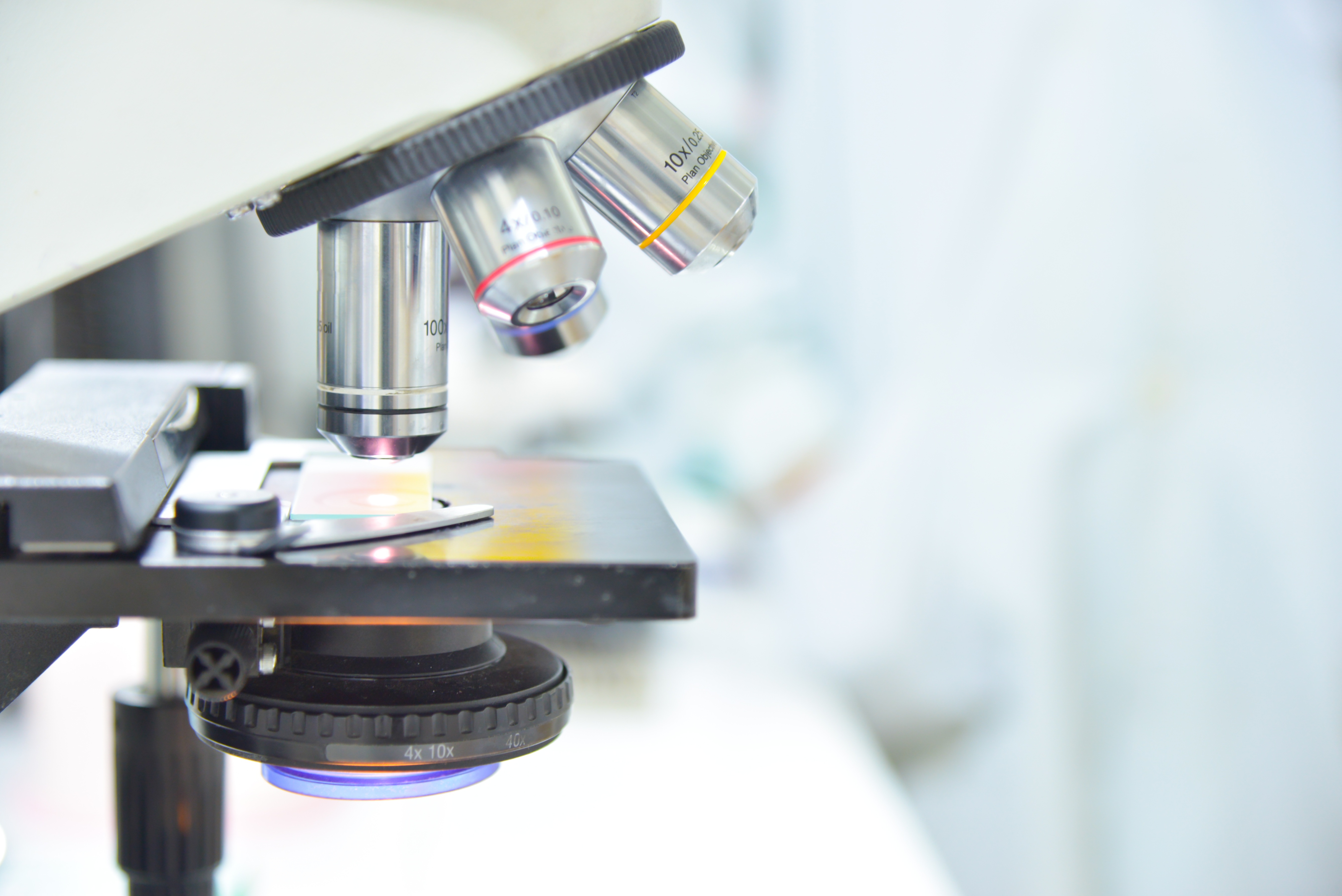 Close-up view of a laboratory microscope focusing on a glass specimen slide positioned on the microscope stage. Multiple objective lenses are visible above the slide, including lenses marked with different magnification levels. The image uses a shallow depth of field, with the microscope and slide in sharp focus while the laboratory background appears softly blurred. Bright, clean lighting and scientific equipment emphasize a research or clinical laboratory setting, representing microscopic analysis, pathology, or biomedical research.