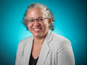Professional head-and-shoulders portrait of a person wearing glasses, a light checkered blazer, and a dark top. The person is facing the camera against a blue gradient studio background. The image is well-lit and framed from the chest up, with the subject positioned slightly angled toward the viewer.