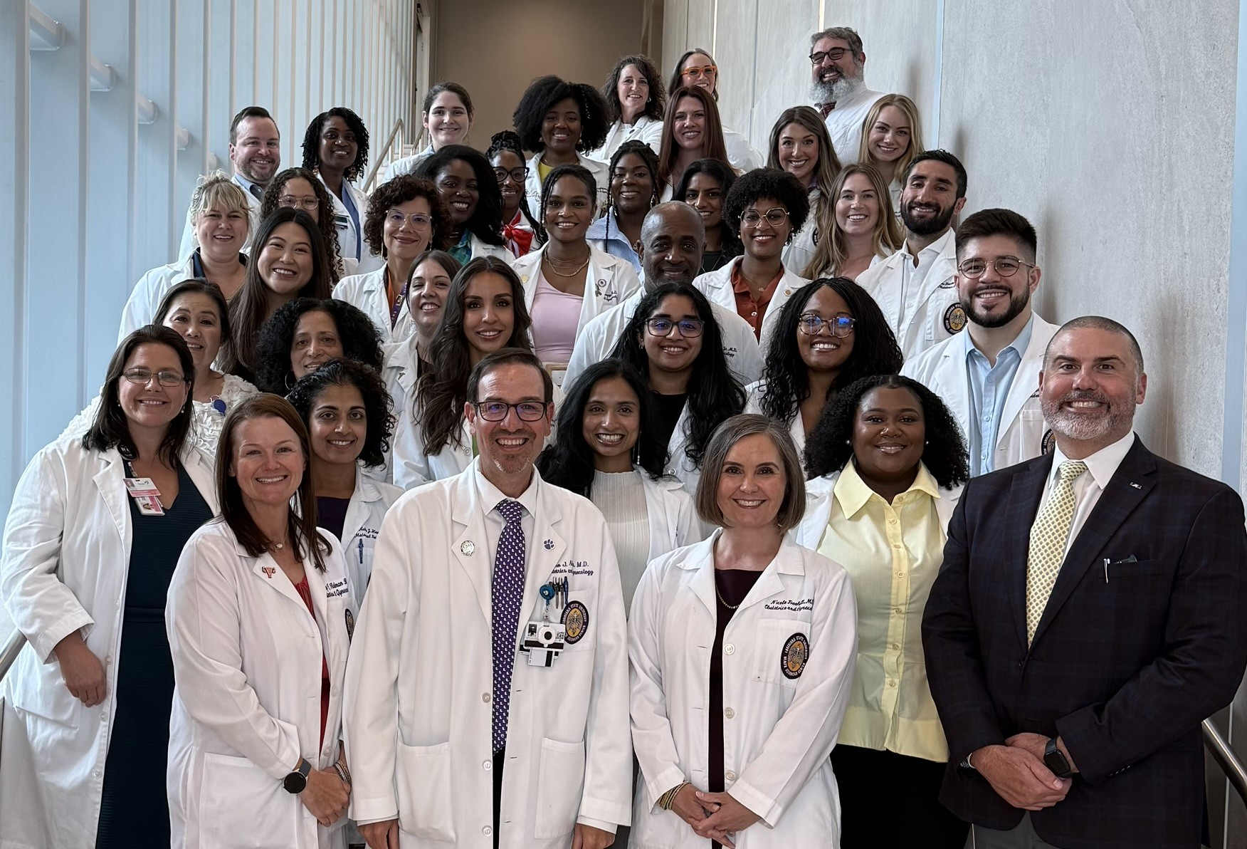 Group portrait of approximately 30 healthcare, research, and academic professionals standing together on an indoor staircase in a bright building with tall windows along one side. Most individuals are wearing white lab coats, while a few are dressed in business attire. The group is arranged in several rows and facing the camera. In the front row, three individuals in white coats stand at the center, with another individual in a dark suit standing to the right. The stairwell features light-colored walls, natural daylight, and metal handrails, creating a formal and professional setting.
