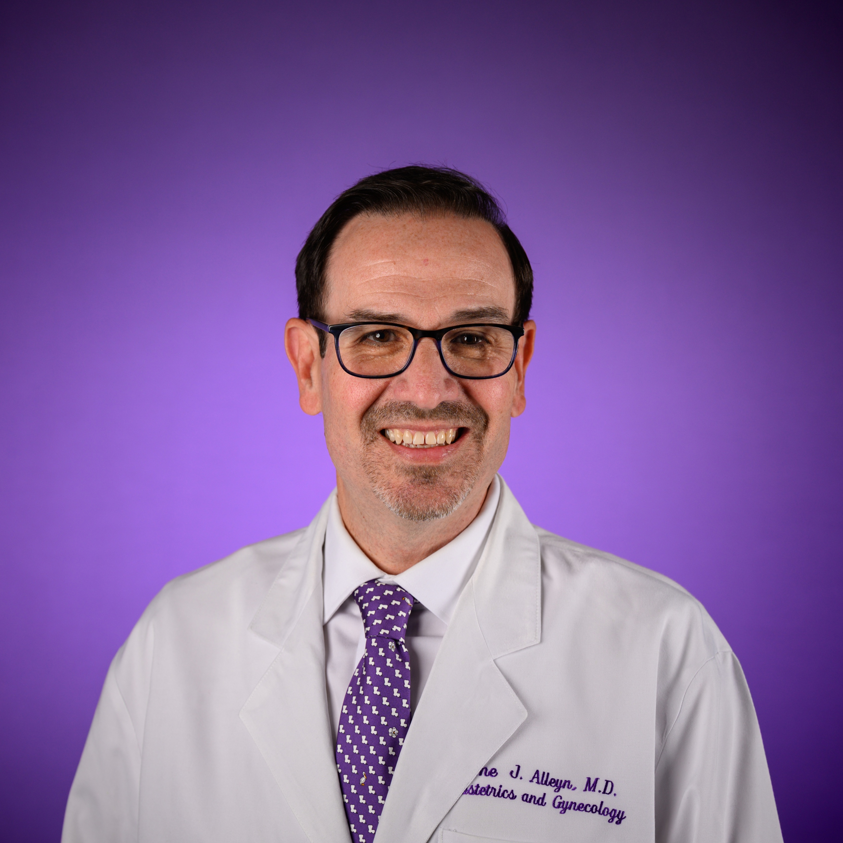 Studio portrait of a person wearing glasses and a white medical lab coat, with a purple patterned tie, standing against a smooth purple gradient background. Embroidered text is visible on the chest of the lab coat.