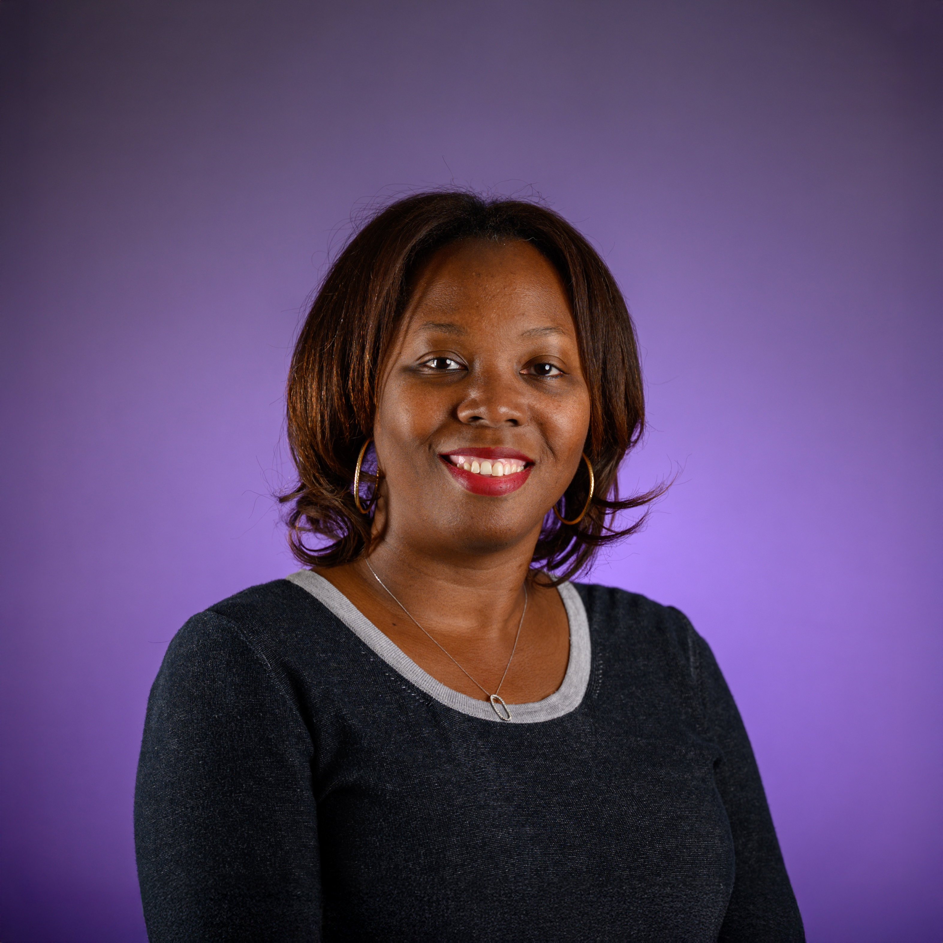 Head-and-shoulders studio portrait of an adult with shoulder-length hair, wearing a dark long-sleeved top with a light trim at the neckline and hoop earrings, facing the camera against a smooth purple background.