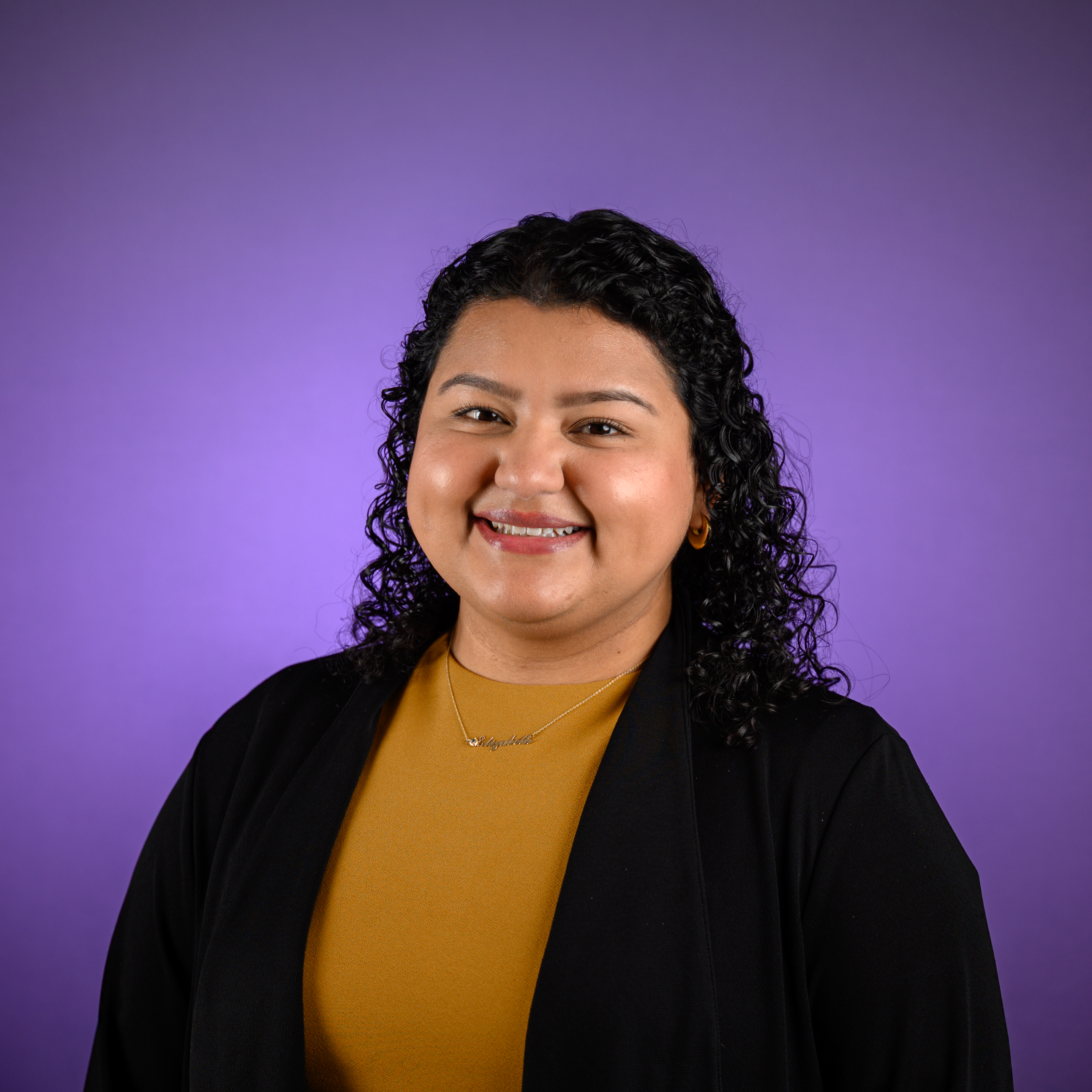 Head-and-shoulders studio portrait of an adult with shoulder-length curly hair, wearing a dark jacket over a mustard-colored top and a small necklace, photographed against a solid purple background.