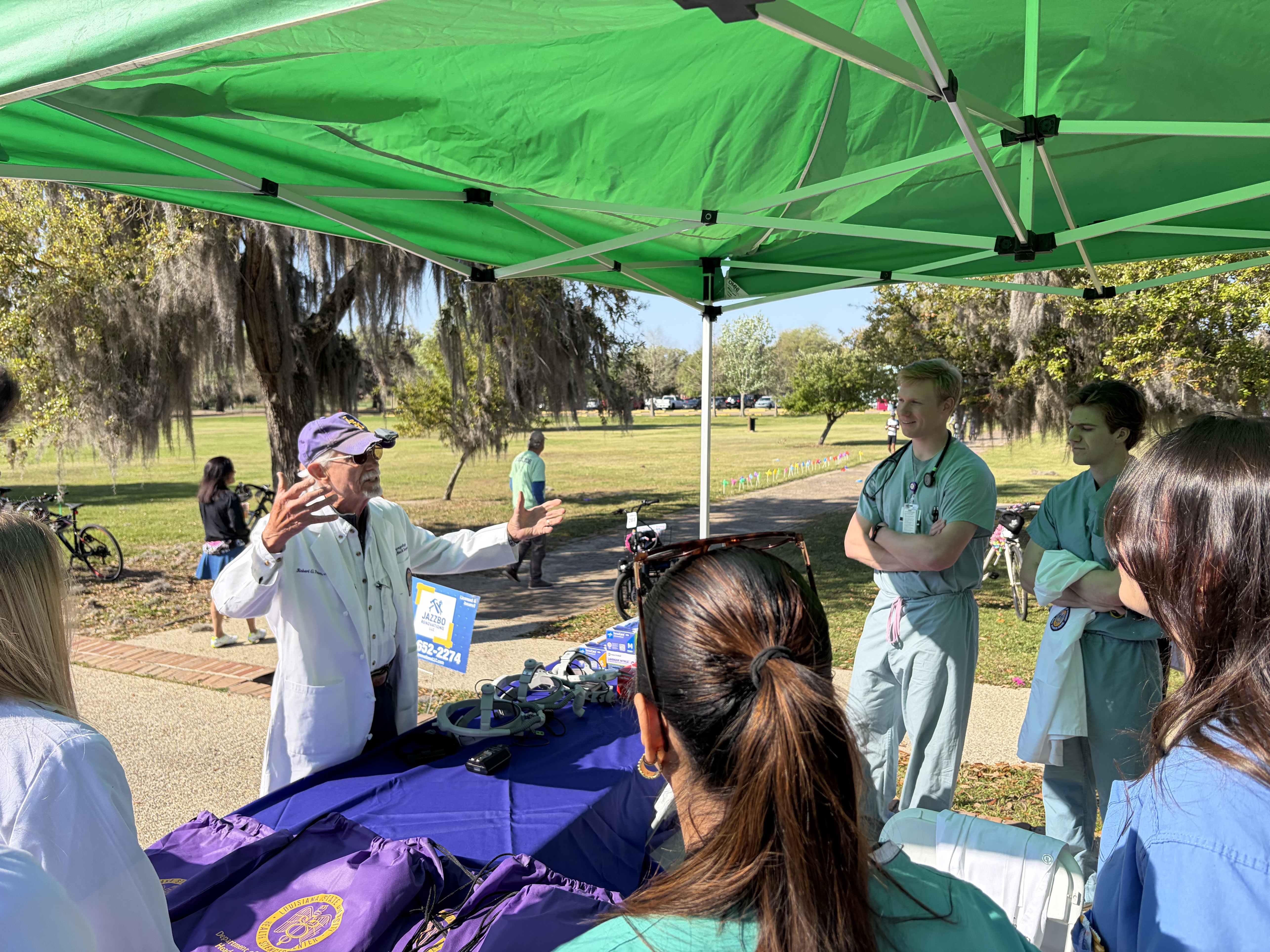 An older man wearing a white coat and purple cap speaks animatedly under a green canopy, gesturing with his hands as he addresses a small group of young adults in medical scrubs gathered around a table with purple materials. The group stands outdoors in a sunny park with grass, trees draped in Spanish moss, and bicycles visible in the background.