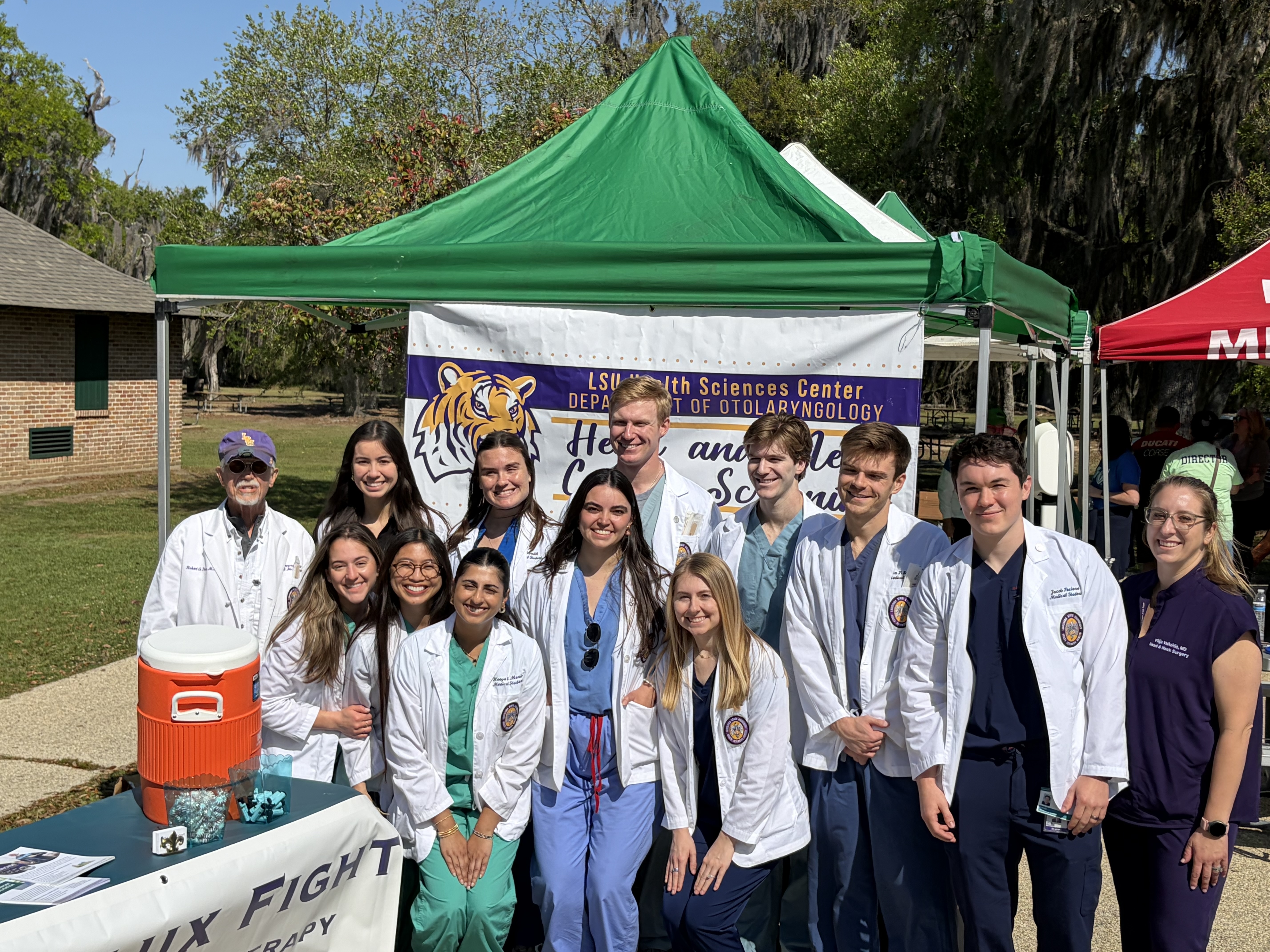 A group of about a dozen young adults and faculty wearing white lab coats and medical scrubs pose for a photo in front of a green canopy tent at an outdoor event. A banner behind them reads “LSU Health Sciences Center Department of Otolaryngology” with a tiger logo. A table with informational materials and an orange drink cooler is in front, and trees and buildings are visible in the sunny background.