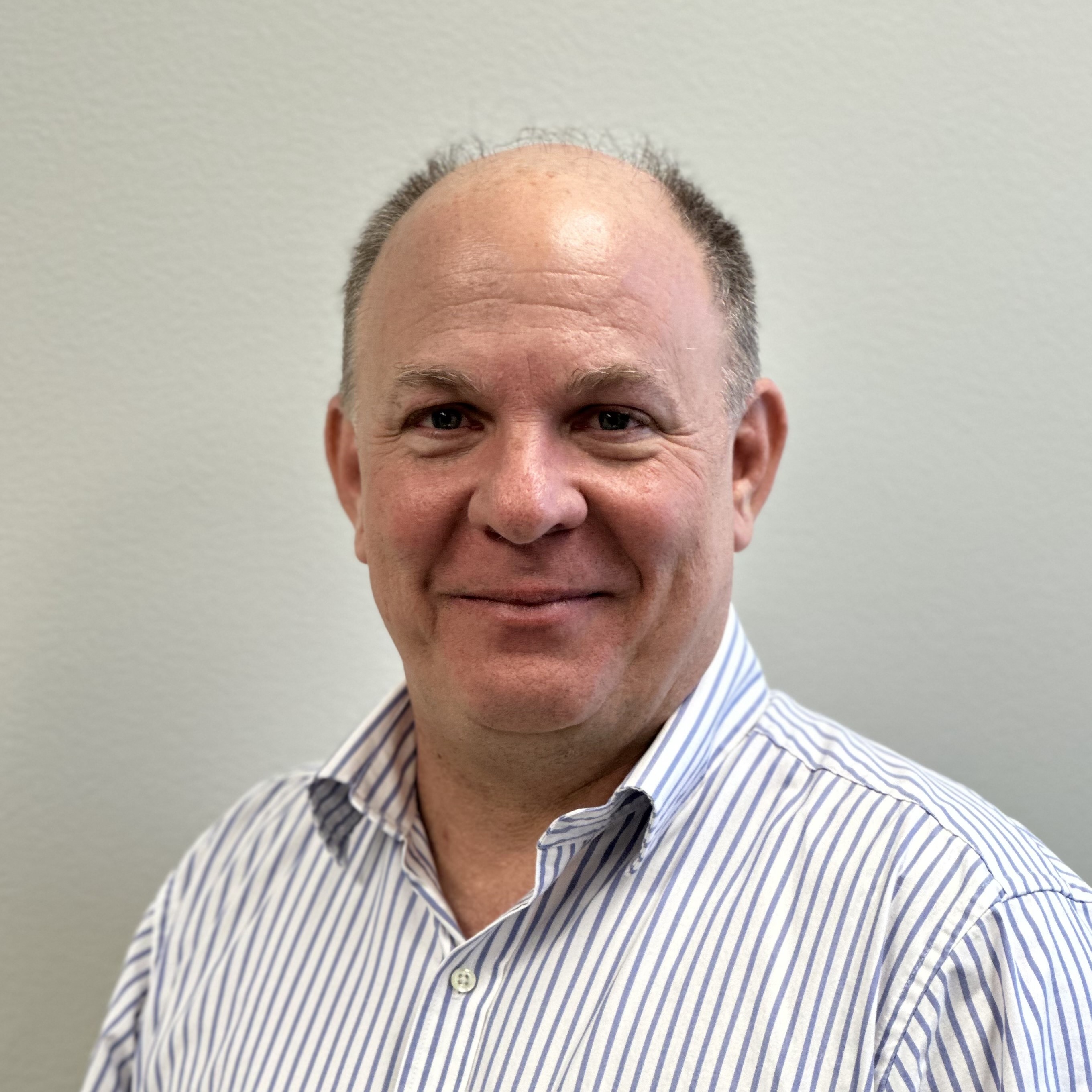 Head-and-shoulders professional portrait of a person wearing a light blue and white striped button-down shirt. The person is facing the camera against a plain light gray background. The image is well lit, with the face in sharp focus and the background softly blurred.