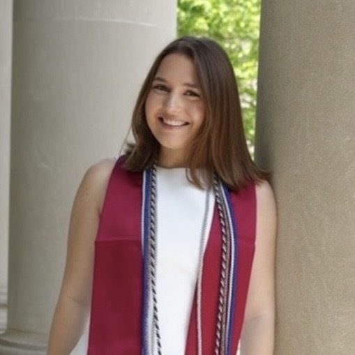 Person standing between large stone columns outdoors, wearing a white sleeveless outfit with a dark red stole and decorative cords draped around the neck. Trees and greenery are visible in the background, and the person is facing the camera.