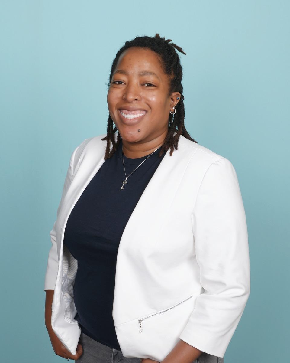 Studio portrait of a person standing against a solid light blue background, facing forward and looking toward the camera. The person is wearing a white blazer over a dark top, along with a necklace and small earrings. The hairstyle consists of short, twisted or loc-style strands pulled back. One hand is placed in a pocket, and the other rests at the side. The lighting is even, clearly illuminating the subject against the simple backdrop.